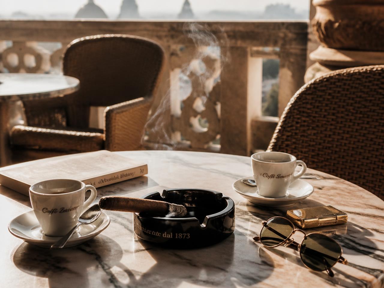 Espresso cups and a lit cigar resting in an ashtray on a sunlit marble terrace table